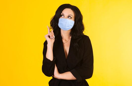 Young businesswoman in mask looks thoughtfully at the top isolated on a yellow background. Coronavirus Outbreak Defense Concept