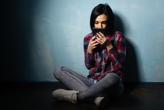 Young sad girl suffering from dependence on social networks sitting on the floor with a smartphone