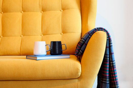 Cup of tea and blue book on a yellow coach with blanket. Still life details in home interior of living room. Cozy home interior, home comfort concept, gender free interior. Modern interior in the room.