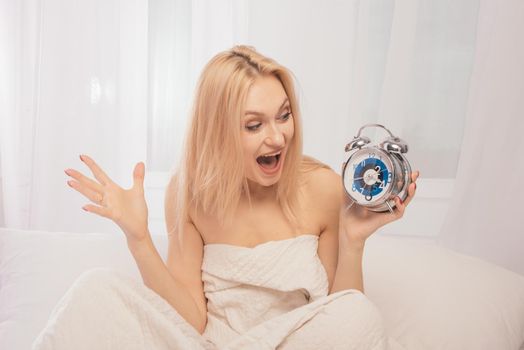 Shocked adult woman in bed with an alarm clock in hand. She screams with surprise that she overslept to work