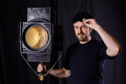 Attractive bearded man hand on the cap stands near a stage spotlight on black background