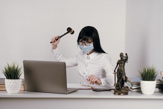 Young woman lawyer or judge works remotely in the office with a protective mask during an epidemic. Swings a hammer to pronounce judgment.