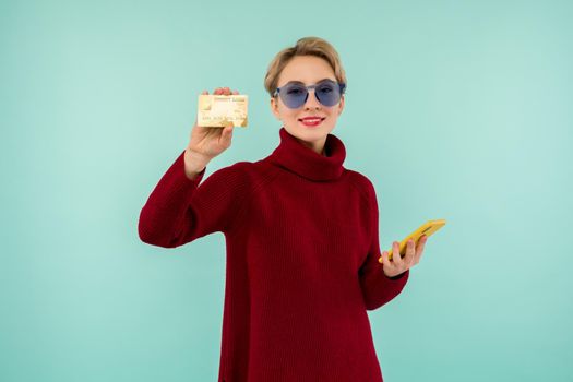 Portrait of beauty young girl in sunglasses showing plastic credit card while holding mobile phone isolated over blue background