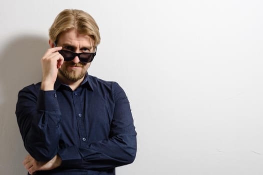 Stylish man in a blue shirt and sunglasses standing near a white wall and a hand holding glasses