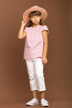 Pretty girl in stylish summer clothes. Child in pink t-shirt, white pants touching her straw hat posing on brown background in studio. Full length portrait of cute girl looking seriously at camera