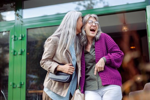 Joyful senior women friends with natural grey hair have fun on modern city street on nice autumn day. Long-time friendship relationship