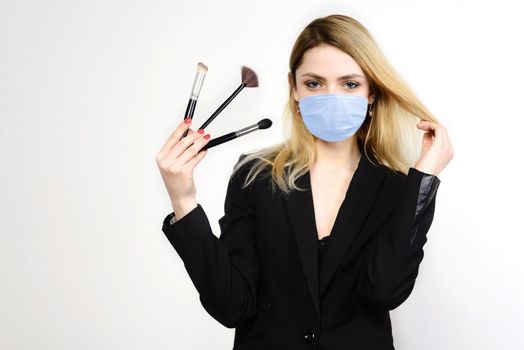 Attractive blonde Make-up artist in mask holding brushes on white background, Coronavirus Outbreak Defense Concept