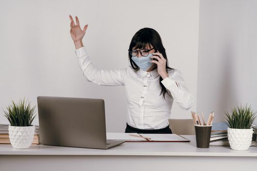 A young woman works remotely in the office with a protective mask during an epidemic. Conducts a video conference and talks on the phone