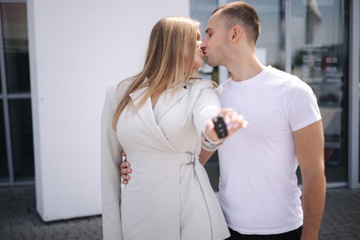 Beautiful young couple kiss each other after baying new car from car showroom. Happy man and woman hold keys.