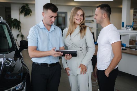 Salesperson talking with couple and help choosing new car. Man and woman in car showroom.