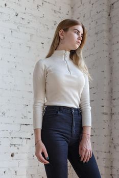 portrait of cute thoughtful teen model wearing white sweater and black jeans. caucasian skinny young female with blond hair stands in front of white brick wall. natural pretty lady standing at studio