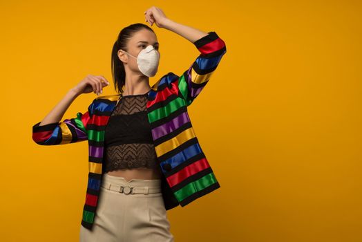 Beautiful slim girl fashion model posing in a protective respirator on a yellow background. LGBT community rainbow color jacket
