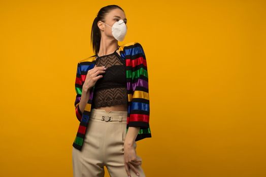 Beautiful slim girl fashion model posing in a protective respirator on a yellow background. LGBT community rainbow color jacket