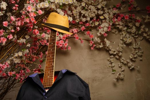 guitar is dressed in a man's blue shirt and a hat with a fretboard near a wall of flowers.