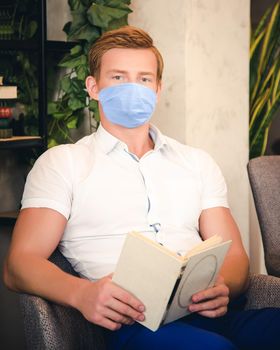 Handsome man in mask reading book self-isolation mode at home is sitting on couch - image