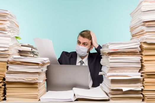 A male accountant or company manager works in an office in a pandemic in view of the accumulated paper work. A protective medical mask is on the face. On the desktop are large stacks of documents. - image