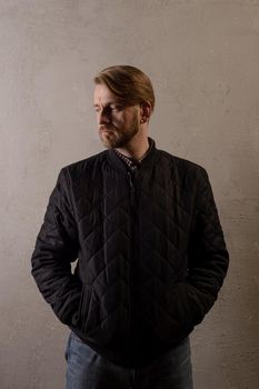 Cute middle-aged bearded man in a black jacket and jeans stands behind the wall looking away. copy space. deadpan without retouching