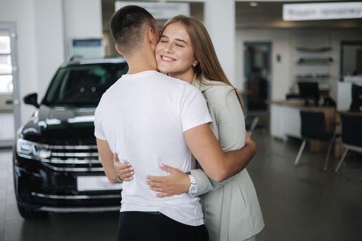 Happy woman hug his husband afrer buying car in car showroom. Man and woman buy new car.