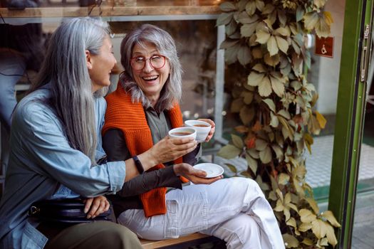 Positive senior Asian woman with friend clink cups of delicious drinks sitting on bench near cozy cafe outdoors. Long-time friendship