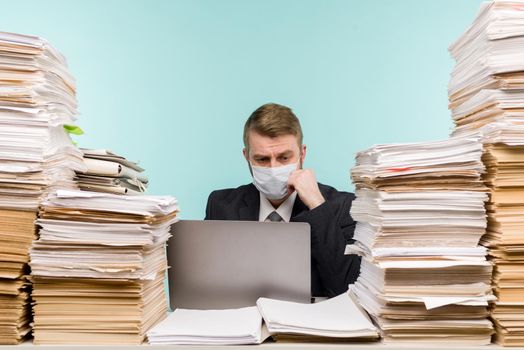 A male accountant or company manager works in an office in a pandemic in view of the accumulated paper work. A protective medical mask is on the face. On the desktop are large stacks of documents. - image