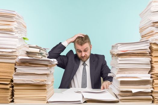 Shocked businessman sitting at the table with many papers in office, he is overloaded with work - image