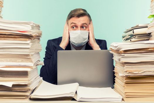 A male accountant or company manager works in an office in a pandemic in view of the accumulated paper work. A protective medical mask is on the face. On the desktop are large stacks of documents. - image