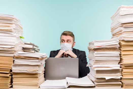 A male accountant or company manager works in an office in a pandemic in view of the accumulated paper work. A protective medical mask is on the face. On the desktop are large stacks of documents. - image