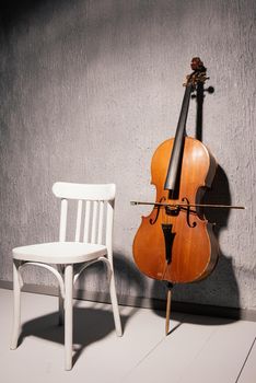 old battered cello and chair standing near a gray textured wall at school or practice room.