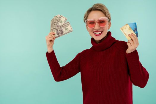 Portrait of happy smiling caucasian woman holding credit card and usa dollars money while looking at camera isolated over blue background.