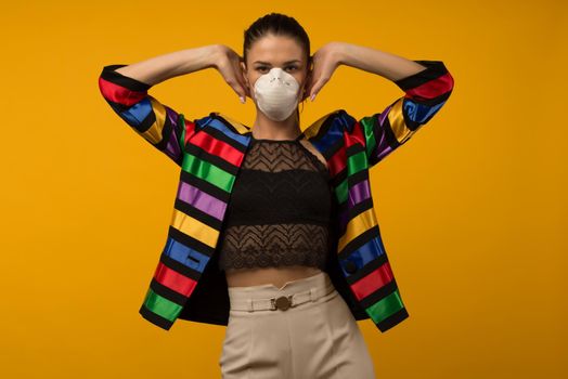 Beautiful slim girl fashion model posing in a protective respirator on a yellow background. LGBT community rainbow color jacket