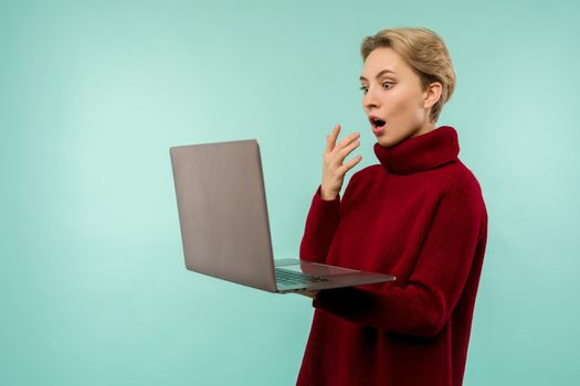 Surprised happy blonde woman in sweater looking laptop computer screen with open mouth over blue background - image