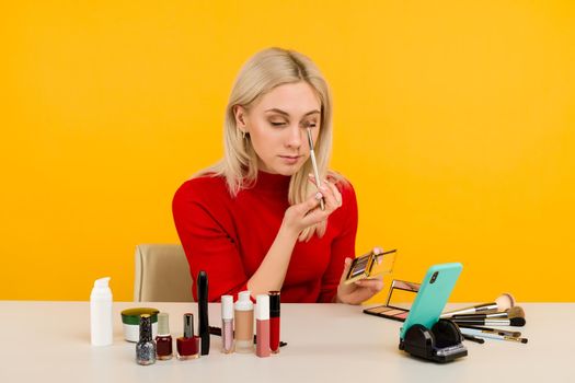 Candid shot of cute young Caucasian woman blogger presenting beauty products and broadcasting live video to social network, using brush to apply eyeshadow while recording everyday make up tutorial