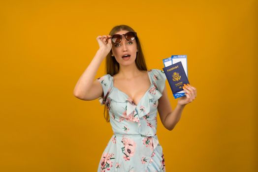 Pop-eyed young woman in blue dress with flowers and sunglasses is holding airline tickets with a passport on a yellow background. Rejoices in the resumption of tourism after the coronovirus pandemic.