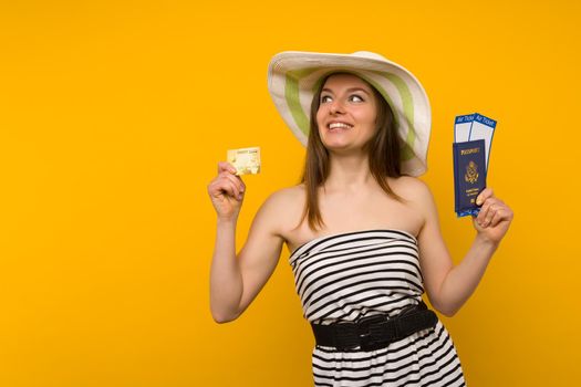 Joyful young woman in a straw hat and a striped dress is holding airline tickets with a passport and a credit card on a yellow background. Rejoices in the resumption of tourism after the coronovirus pandemic.