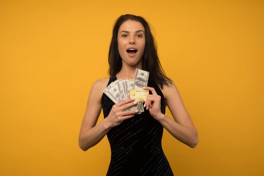 Photo of pleased happy young woman posing isolated over yellow wall background holding money and credit or debt card. - image