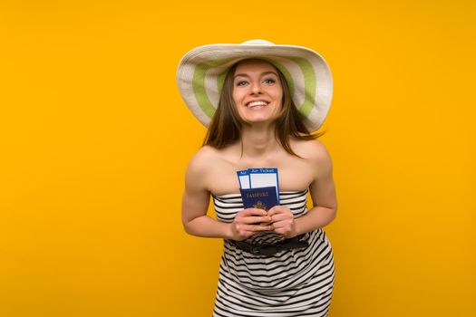 Joyful young woman in a straw hat and a striped dress is holding airline tickets with a passport on a yellow background. Rejoices in the resumption of tourism after the coronovirus pandemic.