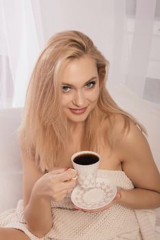 Young woman sitting in bed with a cup of coffie, looking at camera.