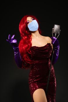 Young romantic redhead woman in mask with very long hair in red dress with microphone on the stand sings with his eyes closed on a black background. Coronavirus Outbreak Defense Concept