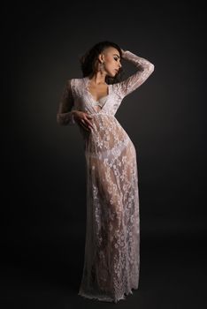 Young slim woman posing in white lace transparent dress on dark background