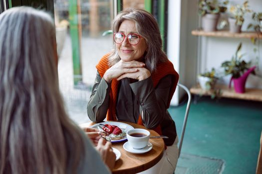 Beautiful grey haired woman with glasses looks at friend sitting at small table in cozy cafe. Long-time friendship