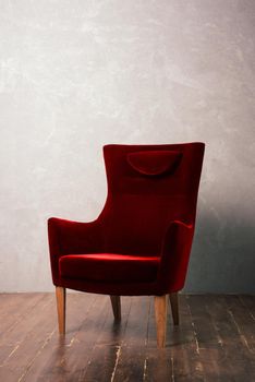 Luxurious red velor armchair stands near the textural wall on a dark wooden floor.