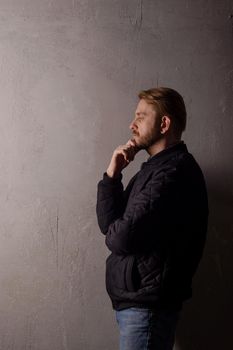 Pensive middle-aged bearded man in a black jacket and jeans stands near the wall looking away. copy space. deadpan without retouching