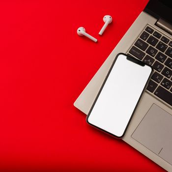 Tula, Russia - May 24,2019: Apple iPhone X and Airpods on red background with notebook. The screen of the smartphone is white. Mockup.