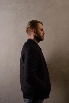 An attractive middle-aged bearded man in a black jacket and jeans stands behind the wall sideways. copy space. deadpan without retouching