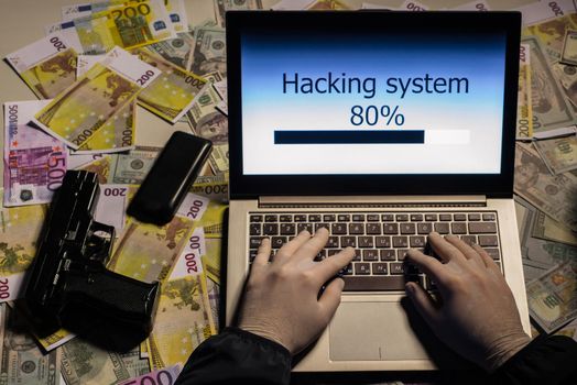 Top view on the hands of an attacker programmer hacking a data server from his laptop. Money is thrown on the table, lies a telephone, a gun, . The concept of cybercrime and hacking database