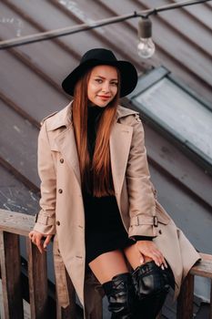 A stylish young woman in a beige coat and black hat sits on a rooftop in the city center. Women's street fashion. Autumn clothing.Urban style.