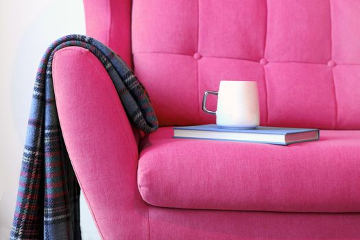 Cup of tea and blue book on a pink coach with blanket. Still life details in home interior of living room. Cozy home interior, home comfort concept, gender free interior modern interior