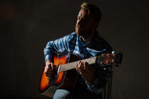 A musician with his eyes closed in a plaid shirt plays fusion on an electroacoustic guitar. Low key portrait