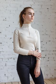 portrait of cute thoughtful teen model wearing white sweater and black jeans. caucasian skinny young female with blond hair stands in front of white brick wall. natural pretty lady standing at studio