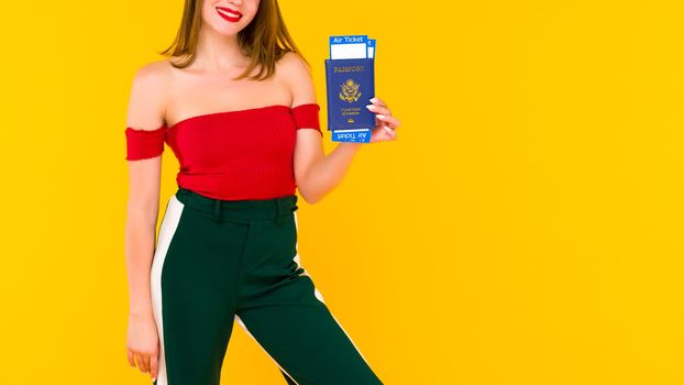 A young woman holds a passport and airline tickets. Focus on a hand with tickets. Stylish girl on in a red blouse and green pants on a yellow background.